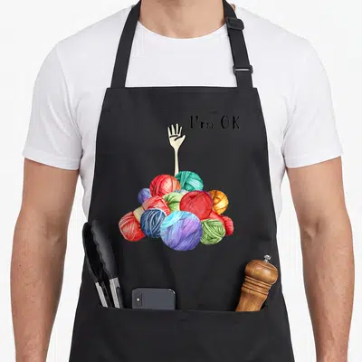 Crochet knitting apron - Hand reaching from red blue green purple and black yarn pile with I'm OK text — shown on a black cotton canvas apron against a white background