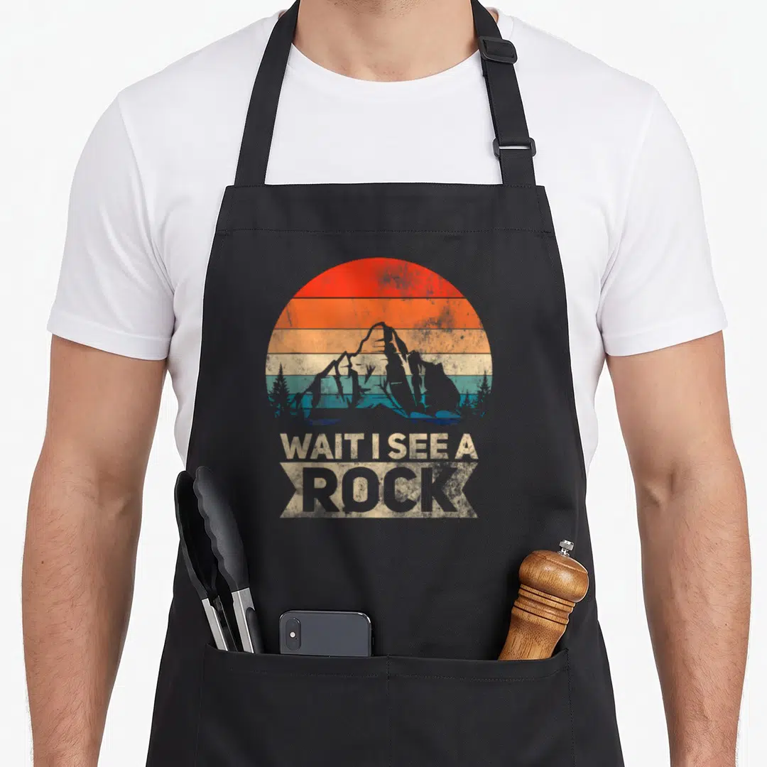 Wait see geologist apron - Retro rock formation with striped sun and text WAIT I SEE A ROCK below — shown on a black cotton canvas apron against a white background