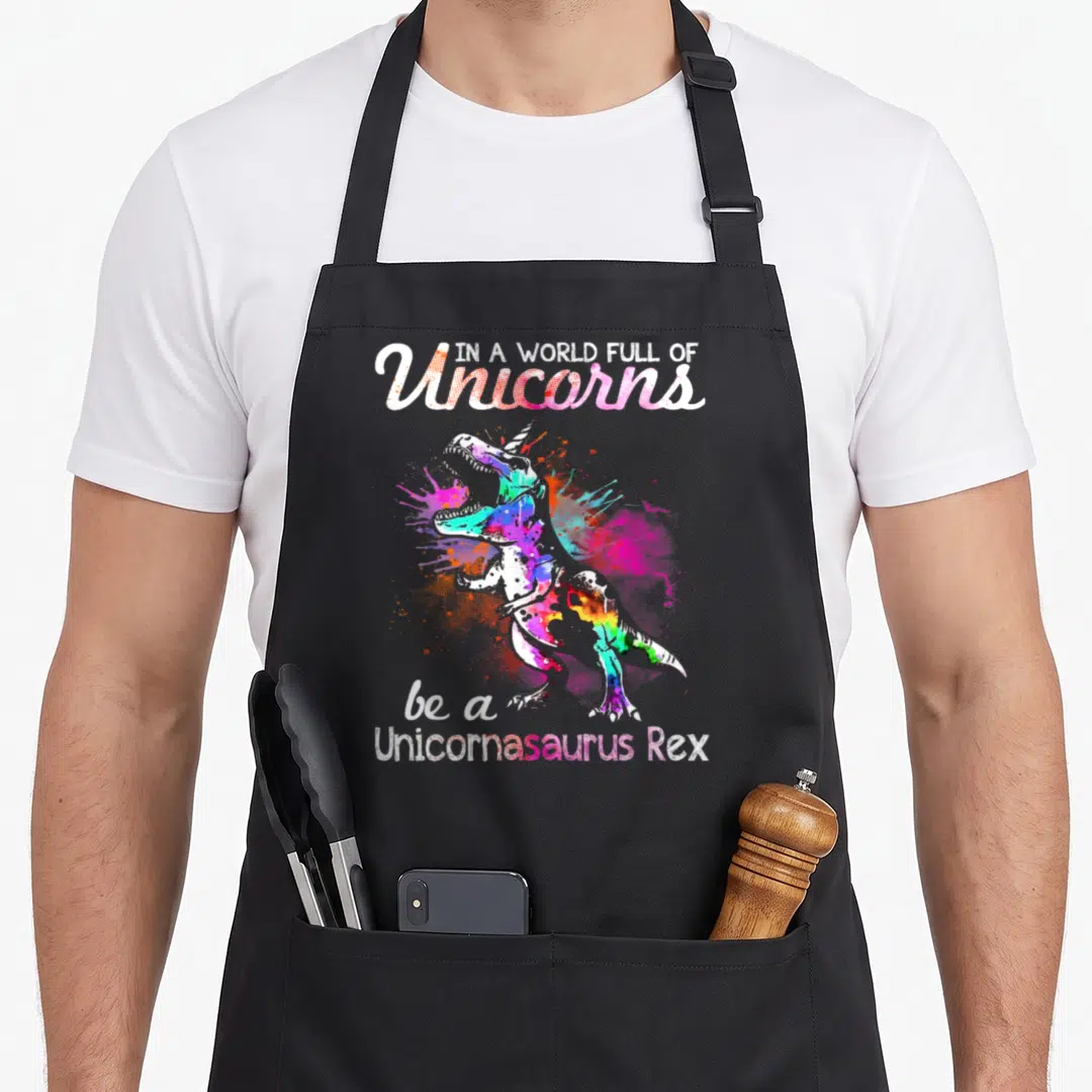 Rex unicorn apron - Watercolor T-Rex with unicorn horn and text be a Unicornasaurus Rex in pink and blue splatters — shown on a black cotton canvas apron against a white background