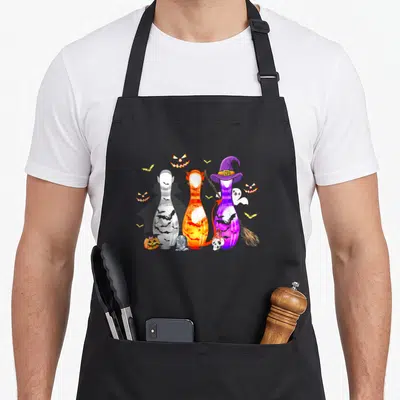 Bowling spooky apron - Three cartoon bowling pins with witch hat, broom, pumpkins, bats, ghost, tombstone, skull — shown on a black cotton canvas apron against a white background
