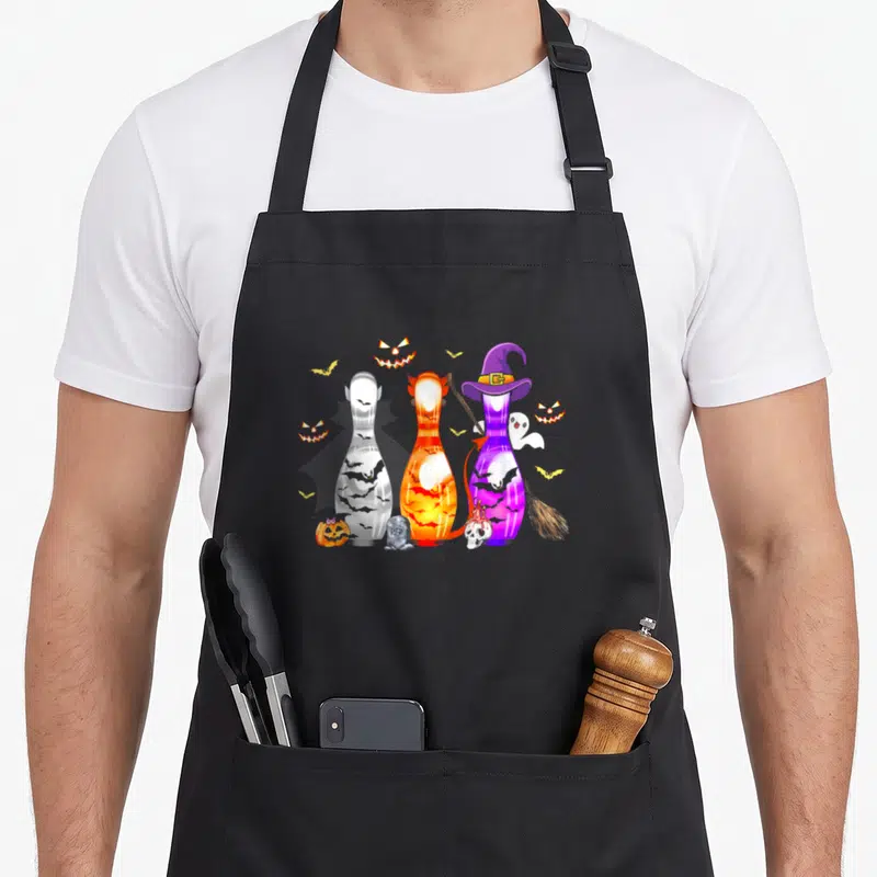 Bowling spooky apron - Three cartoon bowling pins with witch hat, broom, pumpkins, bats, ghost, tombstone, skull — shown on a black cotton canvas apron against a white background