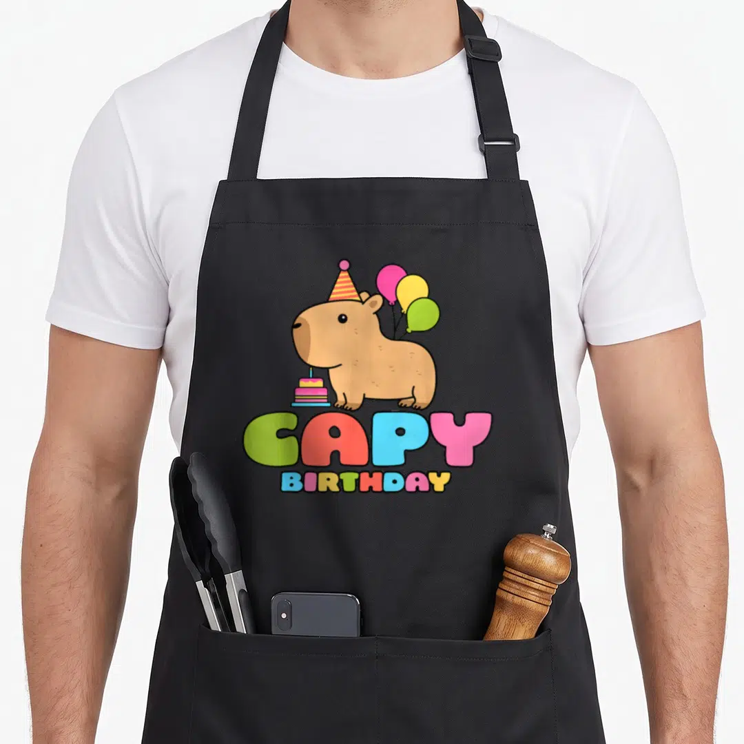 Capy capybara birthday apron - Cartoon brown capybara with party hat, balloons, cake, and colorful CAPY BIRTHDAY text — shown on a black cotton canvas apron against a white background