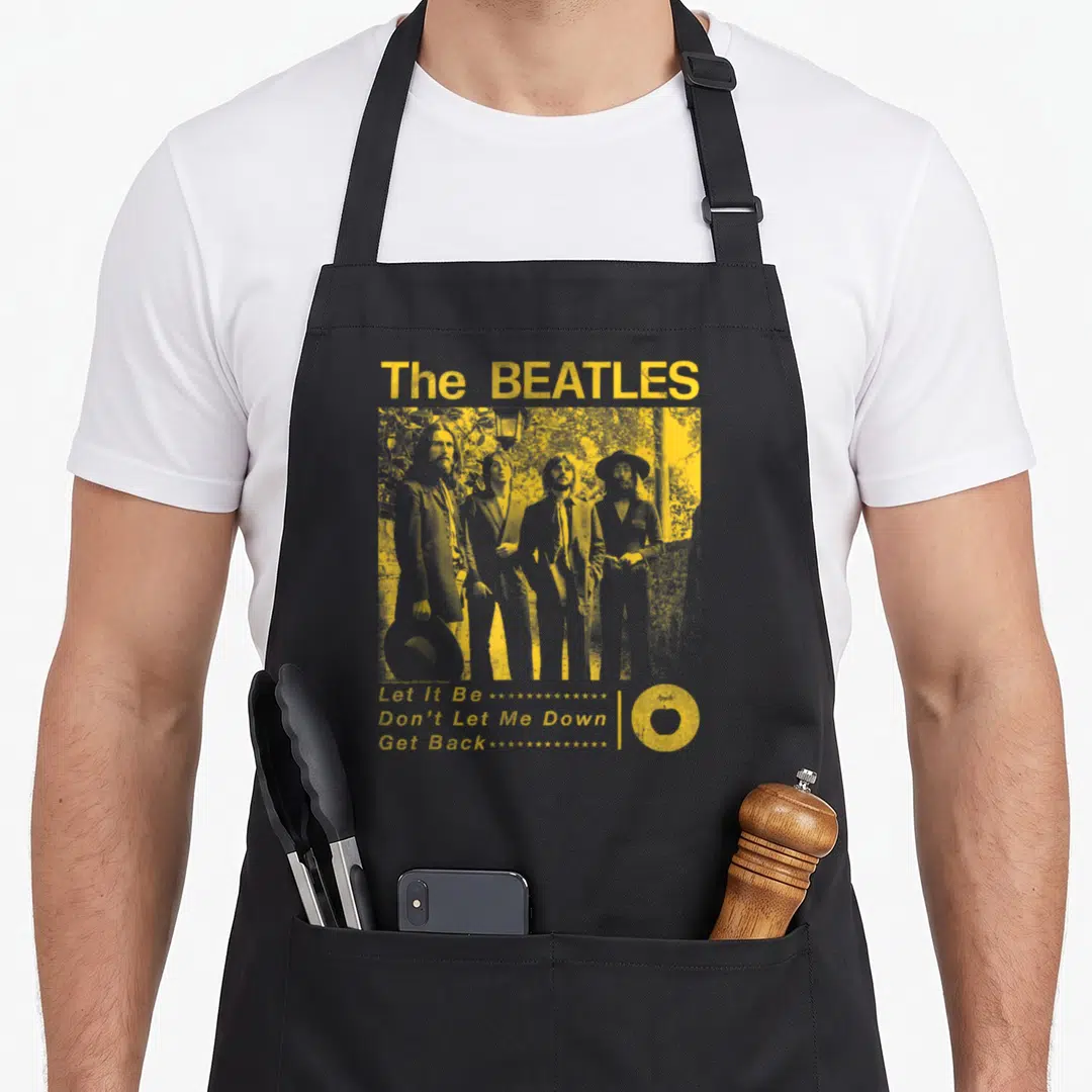 Funny garden apron - Yellow and grey Beatles print with four-member photo, song titles, trailing stars, and Apple logo — shown on a black cotton canvas apron against a white background