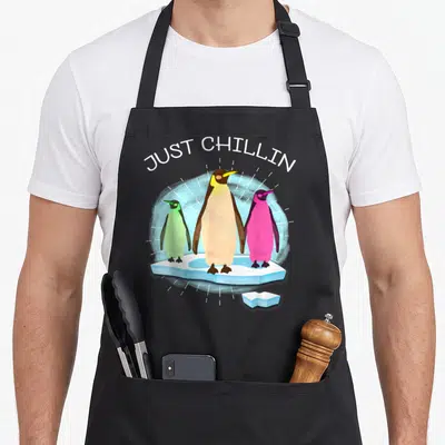 Uab cartoon apron - Three colorful penguins on ice under JUST CHILLIN text — shown on a black cotton canvas apron against a white background