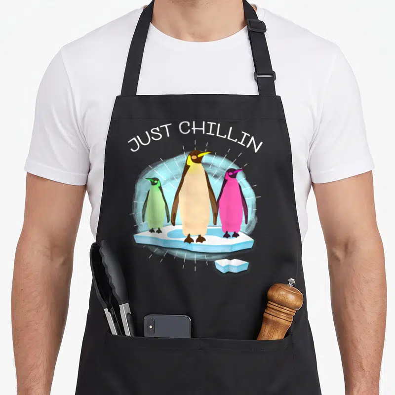 Uab cartoon apron - Three colorful penguins on ice under JUST CHILLIN text — shown on a black cotton canvas apron against a white background