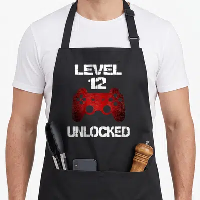 Level unlocked gamer birthday apron - Red distressed game controller with white Level 12 Unlocked text — shown on a black cotton canvas apron against a white background