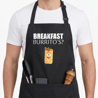 Breakfast mexican apron - Cartoon burrito with face and white text BREAKFAST BURRITO'S? in tan green and red — shown on a black cotton canvas apron against a white background
