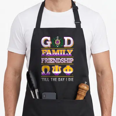 Psi fraternity apron - Vertical text stack GOD FAMILY FRIENDSHIP Omega Psi Phi Greek letters and crest in purple gold — shown on a black cotton canvas apron against a white background