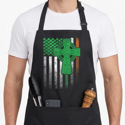 Patrick ireland apron - Green Celtic cross over white and orange stripes with shamrock cluster distressed print — shown on a black cotton canvas apron against a white background