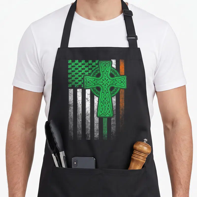 Patrick ireland apron - Green Celtic cross over white and orange stripes with shamrock cluster distressed print — shown on a black cotton canvas apron against a white background