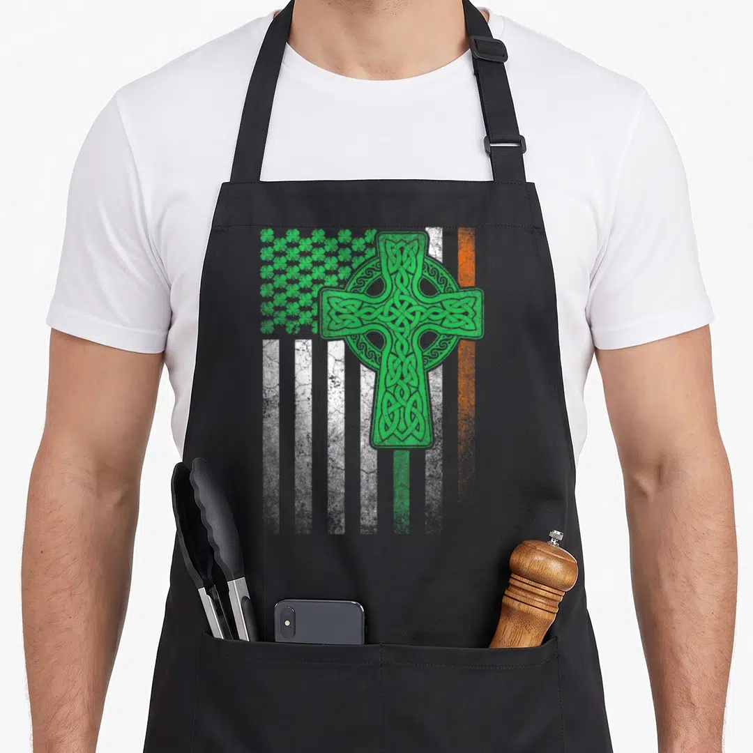 Patrick ireland apron - Green Celtic cross over white and orange stripes with shamrock cluster distressed print — shown on a black cotton canvas apron against a white background