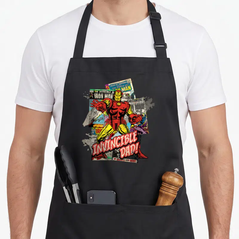 Funny comic father day apron - Iron Man centered over comic pages with red speech bubble text reading Invincible Dad — shown on a black cotton canvas apron against a white background