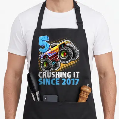Crushing truck aprons - Cartoon monster truck graphic with number 5 and text CRUSHING IT SINCE 2017 in yellow and blue — shown on a black cotton canvas apron against a white background