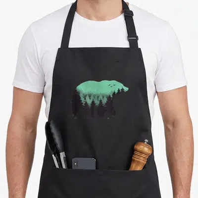 Hunting bear apron - Black bear silhouette filled with pine trees and birds on mint green background — shown on a black cotton canvas apron against a white background