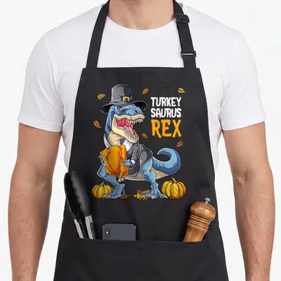 Dinosaur pilgrim apron - Blue T-Rex in pilgrim hat holding roasted turkey with TURKEY SAURUS REX text — shown on a black cotton canvas apron against a white background