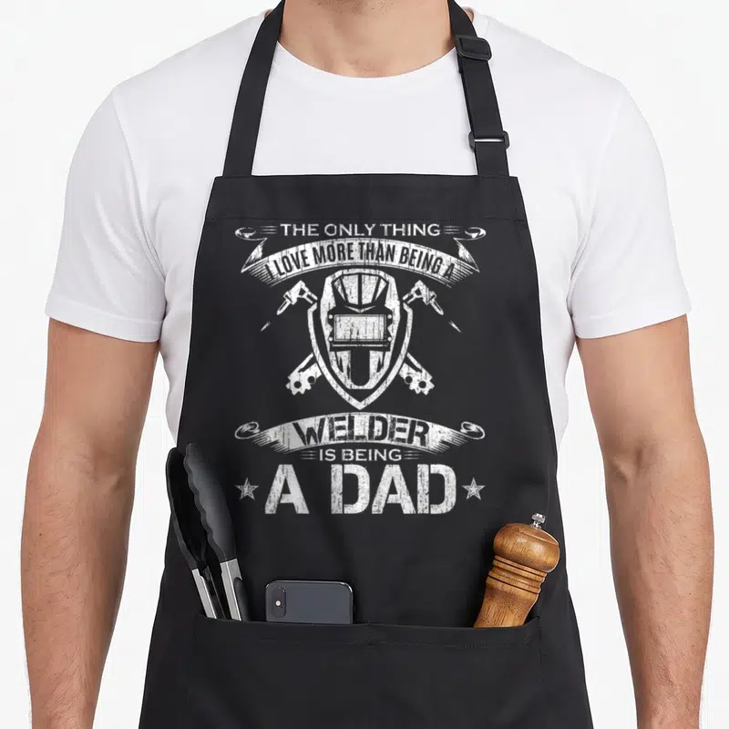 Funny welding welder dad apron - White distressed welding helmet and crossed torches with arched banner text — shown on a black cotton canvas apron against a white background