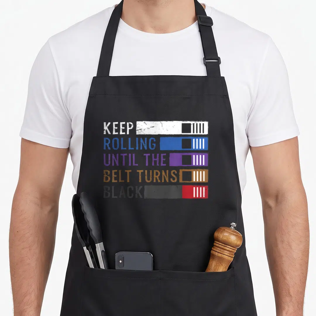 Belt jitsu apron - Keep Rolling Until The Belt Turns Black text with stacked white blue purple brown and black belt — shown on a black cotton canvas apron against a white background