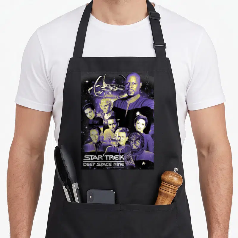 Ds9 crew apron - Photo collage of Deep Space Nine cast members with space station and stacked title text — shown on a black cotton canvas apron against a white background
