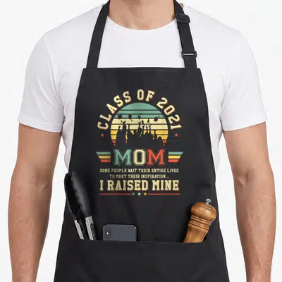 Graduate senior apron - Retro sunburst with graduate silhouettes and text Class of 2021 Mom I Raised Mine — shown on a black cotton canvas apron against a white background