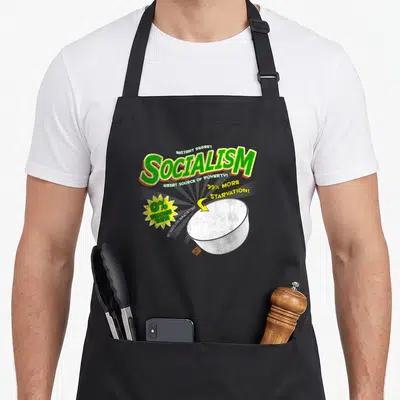 Socialism box apron - Green SOCIALISM text, empty white bowl, yellow starburst, and sarcastic slogans on a cartoon — shown on a black cotton canvas apron against a white background