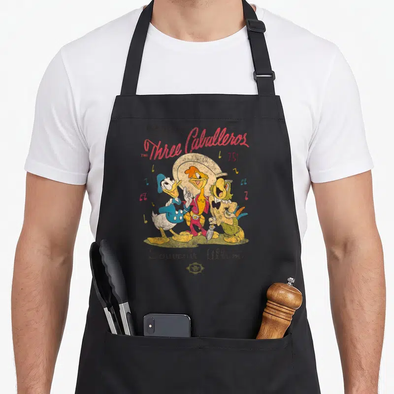 Three novelty apron - Retro Three Caballeros illustration with red script, musical notes, and Souvenir Album text — shown on a black cotton canvas apron against a white background