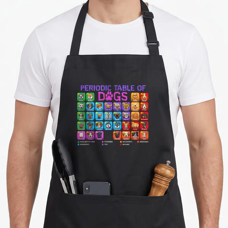 Periodic custom pet apron - Colorful grid of dog heads in purple green blue orange and red squares with breed names — shown on a black cotton canvas apron against a white background