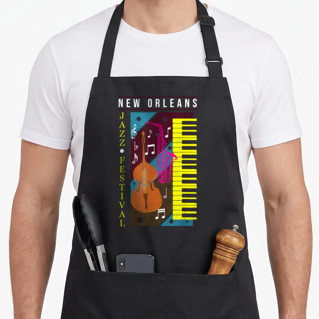 Jazz new orleans apron - Retro cello, pink sax, yellow piano keys, and vertical jazz festival text in blue and brown — shown on a black cotton canvas apron against a white background