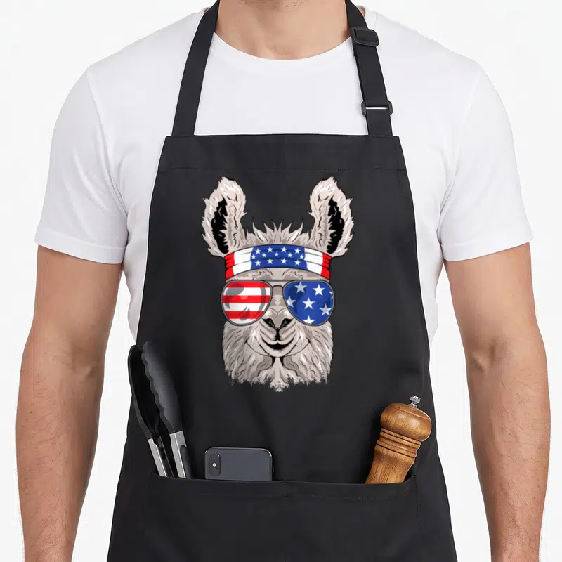 Patriotic alpaca apron - Cartoon llama wearing USA flag sunglasses and bandana in grey, white, red, and blue — shown on a black cotton canvas apron against a white background