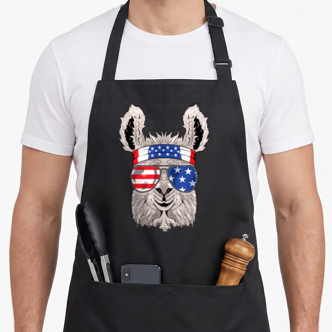Patriotic alpaca apron - Cartoon llama wearing USA flag sunglasses and bandana in grey, white, red, and blue — shown on a black cotton canvas apron against a white background