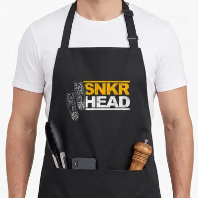 Sneaker shoe apron - Two white sneaker soles beside stacked SNKR HEAD text in distressed yellow and white — shown on a black cotton canvas apron against a white background