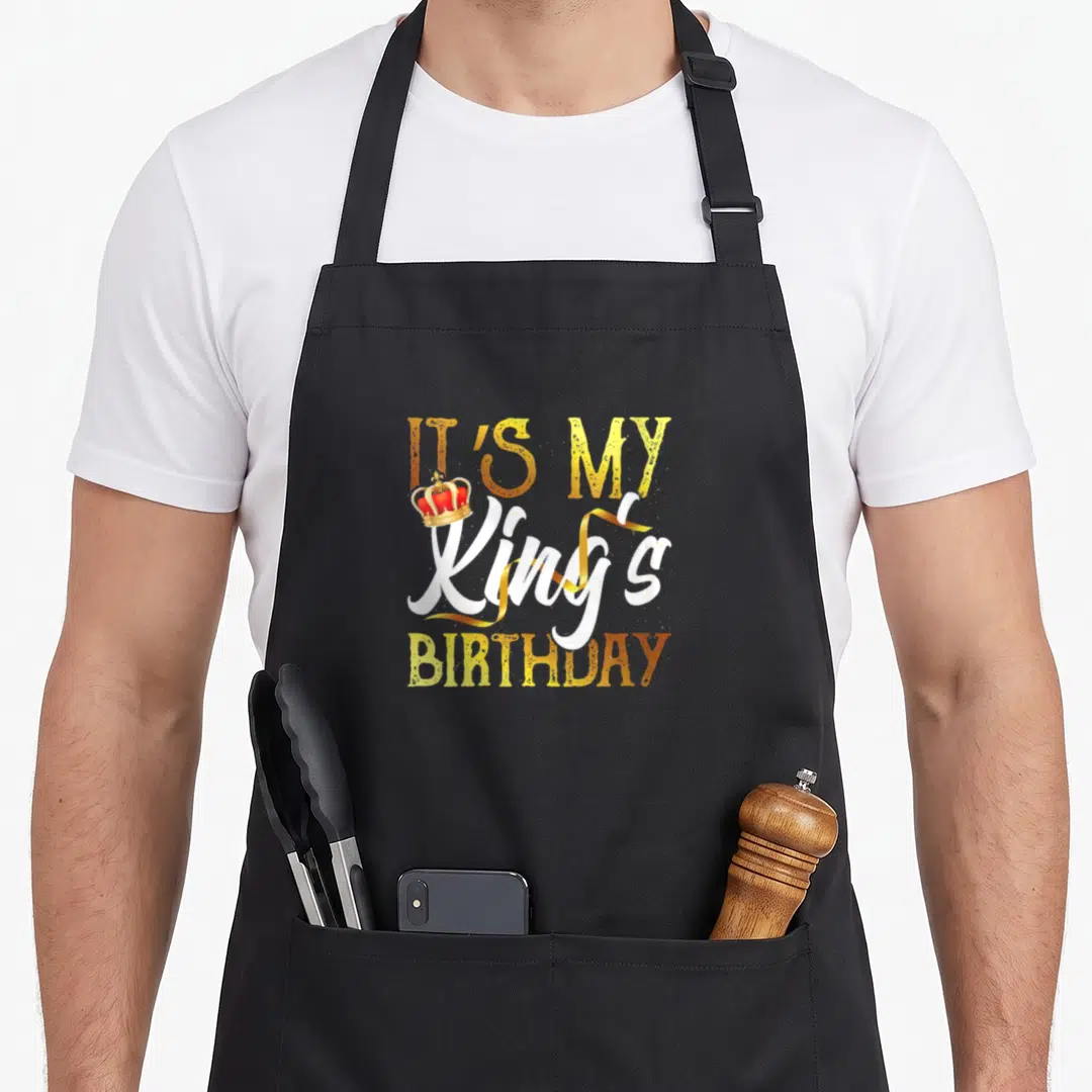 King queen of baking apron - Stacked IT'S MY King's BIRTHDAY text with red crown on K and curling gold ribbon — shown on a black cotton canvas apron against a white background