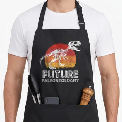 Paleontologist rex dinosaur apron - Distressed T-Rex skeleton over red orange yellow striped sun with future — shown on a black cotton canvas apron against a white background