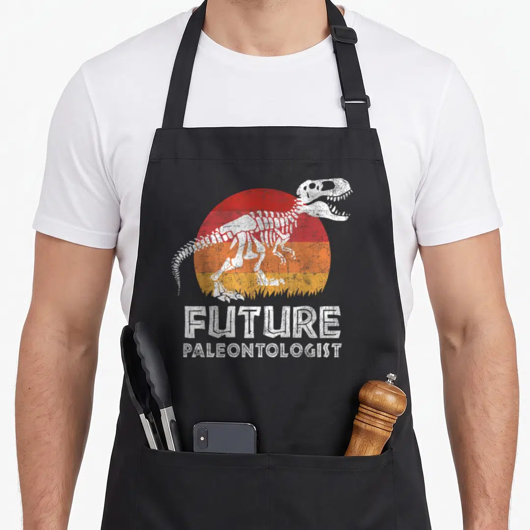 Paleontologist rex dinosaur apron - Distressed T-Rex skeleton over red orange yellow striped sun with future — shown on a black cotton canvas apron against a white background