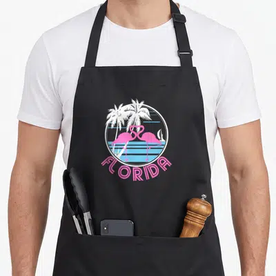 Florida pink flamingo apron - Two pink flamingos forming a heart inside a circle with palm trees and blue stripes — shown on a black cotton canvas apron against a white background
