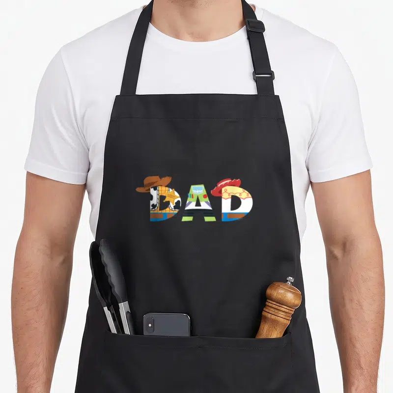 Birthday father day apron - DAD text formed by Woody Buzz and Jessie character illustrations with hats in cartoon style — shown on a black cotton canvas apron against a white background
