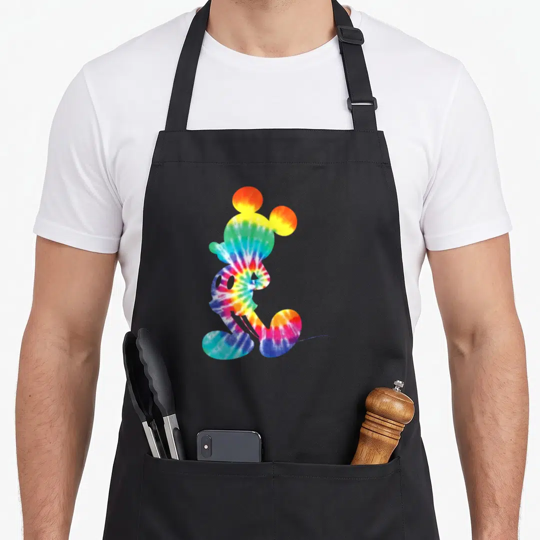 Rainbow tie apron - Mickey Mouse silhouette in tie-dye red yellow green blue purple — shown on a black cotton canvas apron against a white background