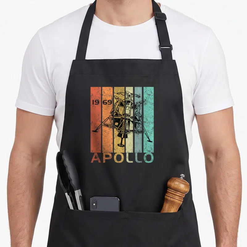 50th anniversary apollo apron - Black lunar module over five orange yellow teal stripes with 1969 date and APOLLO text — shown on a black cotton canvas apron against a white background