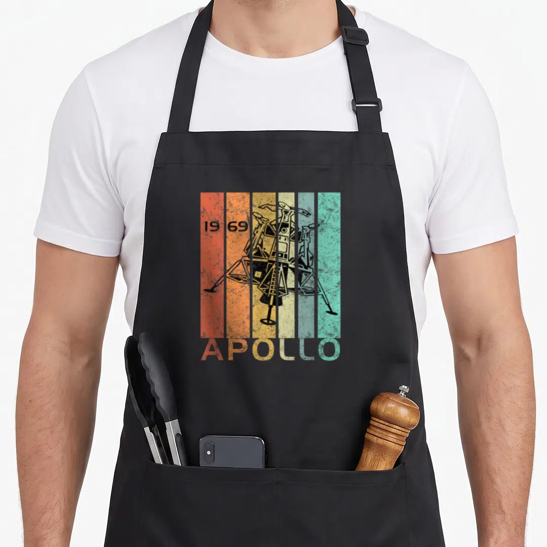50th anniversary apollo apron - Black lunar module over five orange yellow teal stripes with 1969 date and APOLLO text — shown on a black cotton canvas apron against a white background