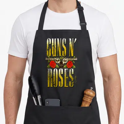 Funny rose gun apron - Crossed silver revolvers wrapped in barbed wire flanked by red roses under distressed yellow text — shown on a black cotton canvas apron against a white background