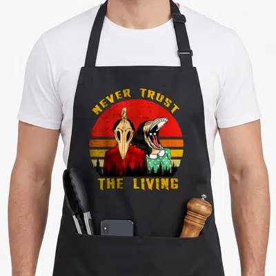 Trust goth apron - Two skeletal figures under NEVER TRUST THE LIVING text against red orange yellow sunset — shown on a black cotton canvas apron against a white background