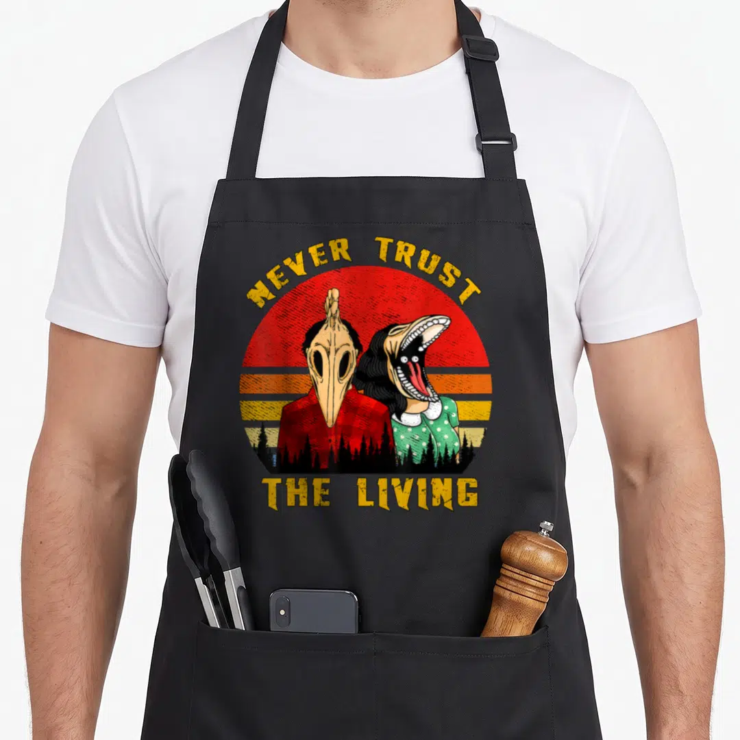 Trust goth apron - Two skeletal figures under NEVER TRUST THE LIVING text against red orange yellow sunset — shown on a black cotton canvas apron against a white background