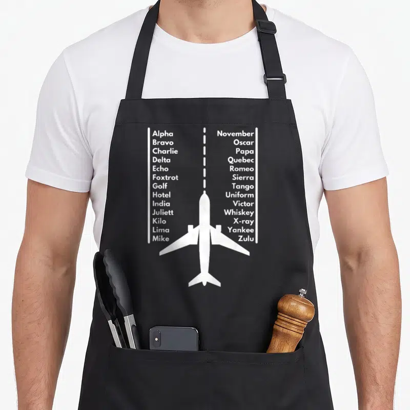 Phonetica airplane apron - White airplane silhouette with dashed flight line and two columns of phonetic alphabet text — shown on a black cotton canvas apron against a white background