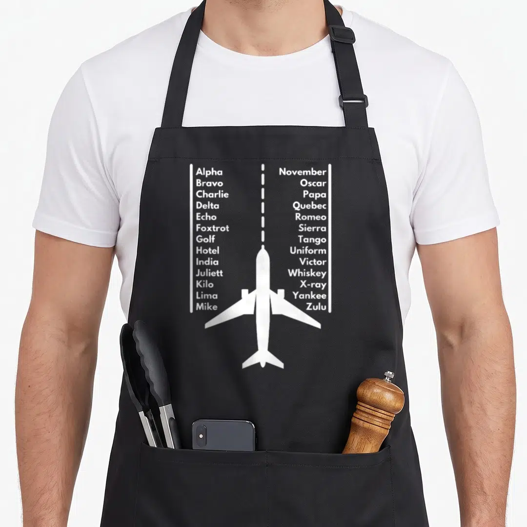 Phonetica airplane apron - White airplane silhouette with dashed flight line and two columns of phonetic alphabet text — shown on a black cotton canvas apron against a white background