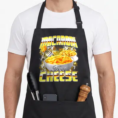 Macaroni meme apron - Arched MACARONI And CHEESE text with two pasta bowls and lightning bolts — shown on a black cotton canvas apron against a white background