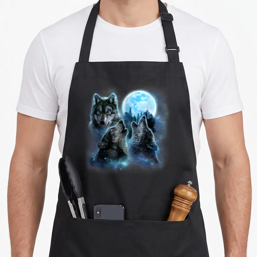 Howling wolves apron - Realistic wolf head and two howling wolves under a blue moon with forest silhouette — shown on a black cotton canvas apron against a white background