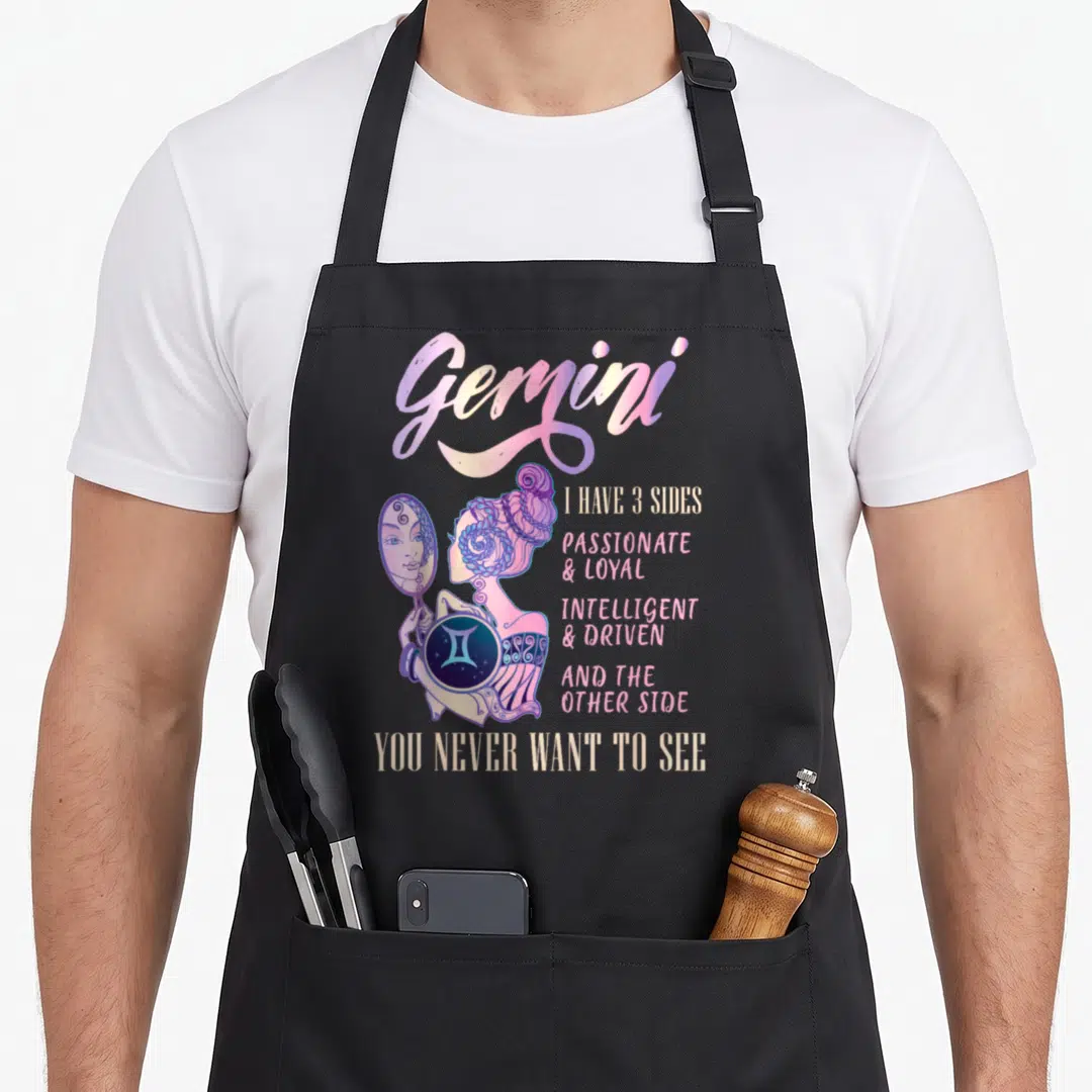 Cute gemini zodiac aprons - Woman holding mirror with Gemini text listing three sides and a final warning in purple — shown on a black cotton canvas apron against a white background
