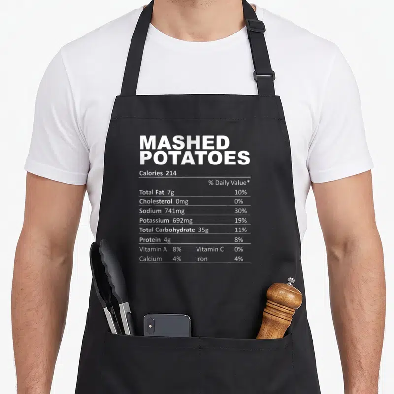 Mashed food aprons - White nutrition label text for Mashed Potatoes on black fabric showing 214 calories and nutrient — shown on a black cotton canvas apron against a white background