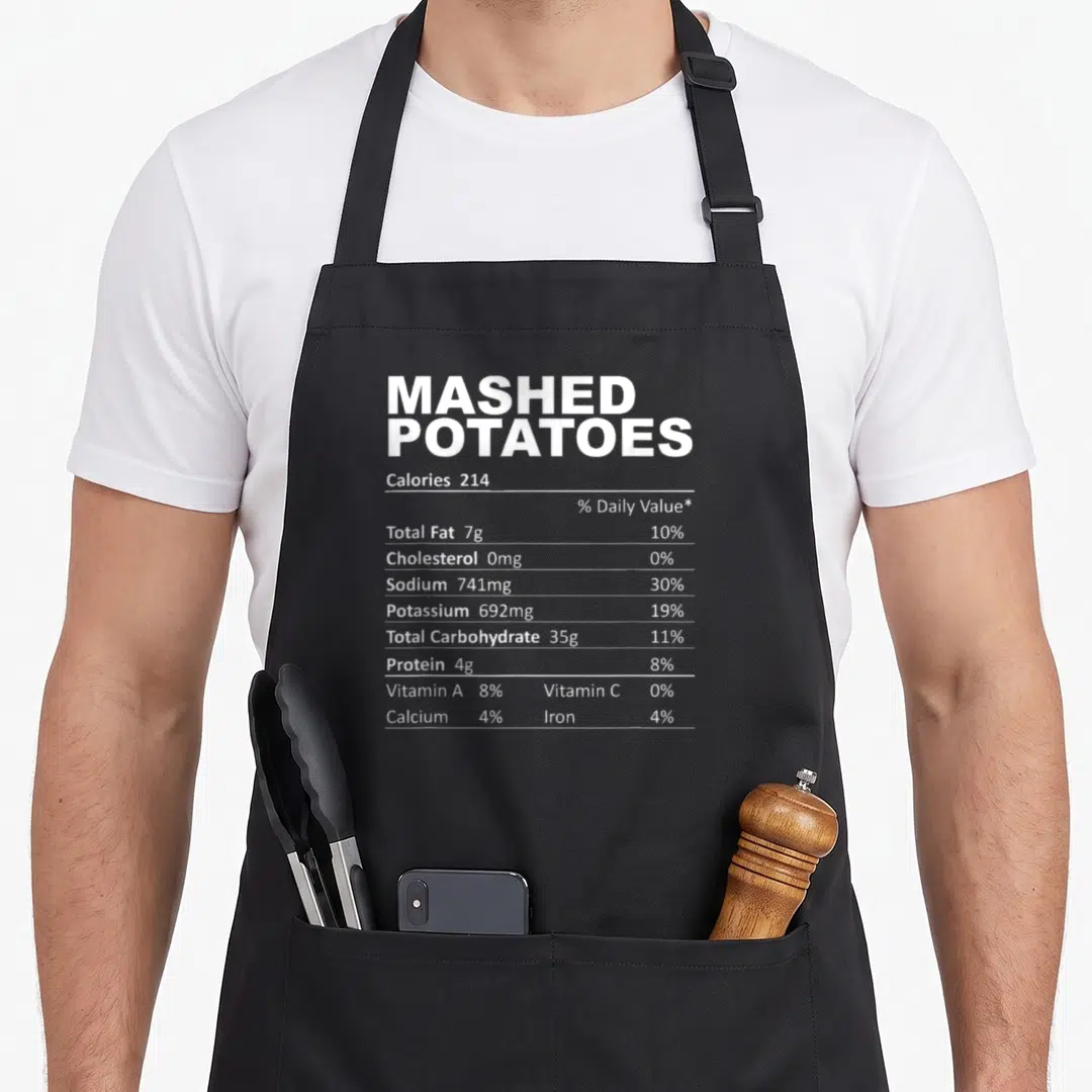 Mashed food aprons - White nutrition label text for Mashed Potatoes on black fabric showing 214 calories and nutrient — shown on a black cotton canvas apron against a white background