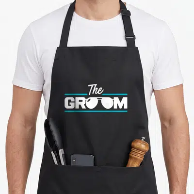 Groomsmen funny wedding aprons - White The GROOM text with glasses lenses replacing letters between teal lines — shown on a black cotton canvas apron against a white background