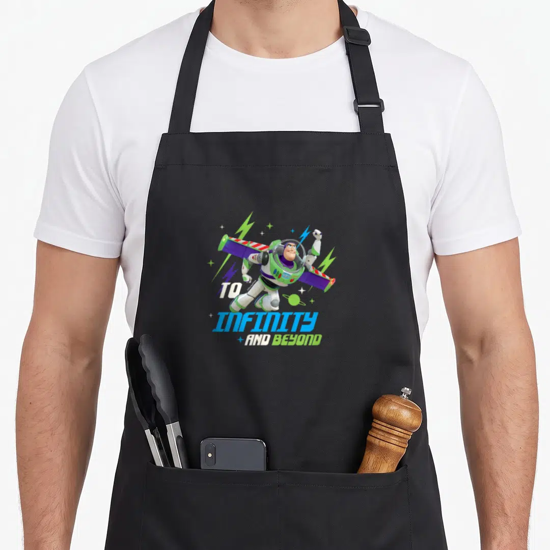 Beyond infinity apron - Buzz Lightyear flying diagonally with TO INFINITY AND BEYOND text in white green blue purple — shown on a black cotton canvas apron against a white background